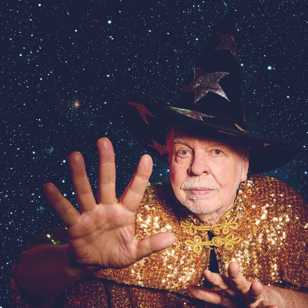 Rick Wakeman wearing a wizard's hat and gold sequence cloak in front of a starry night scene, holding his out towards the camera