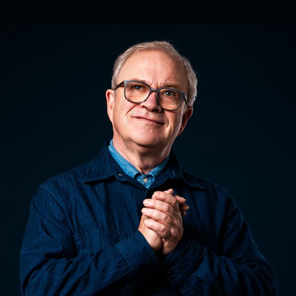 Harry Enfield wearing glasses, looking upwards with his hand clasped