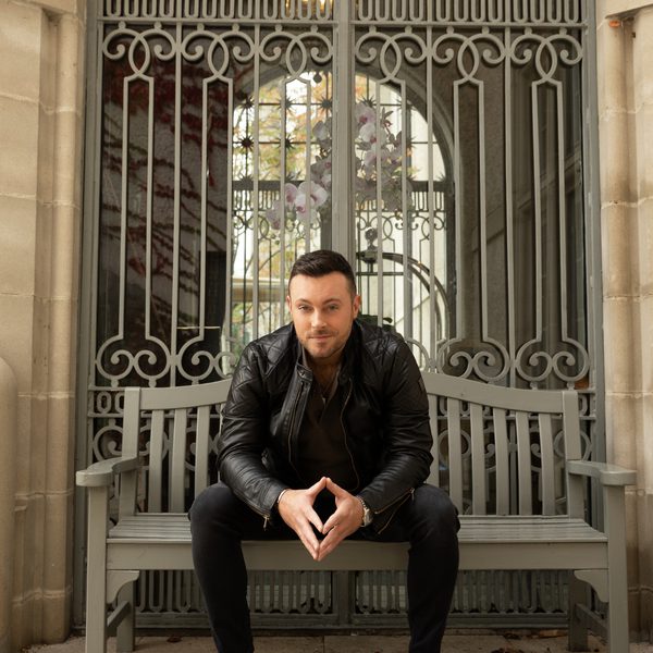 Nathan Carter wearing a black leather jacket and dark clothing sitting on a bench in front of ornate gates
