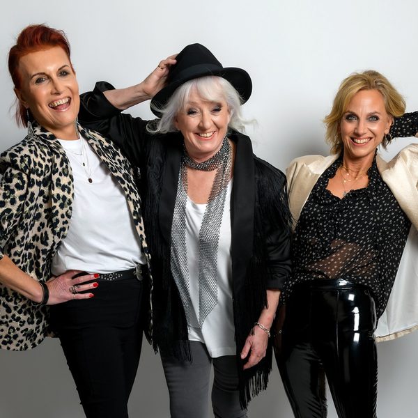 3 middle aged women stood in a line with their hands on their hips wearing bright, shimmery clothing
