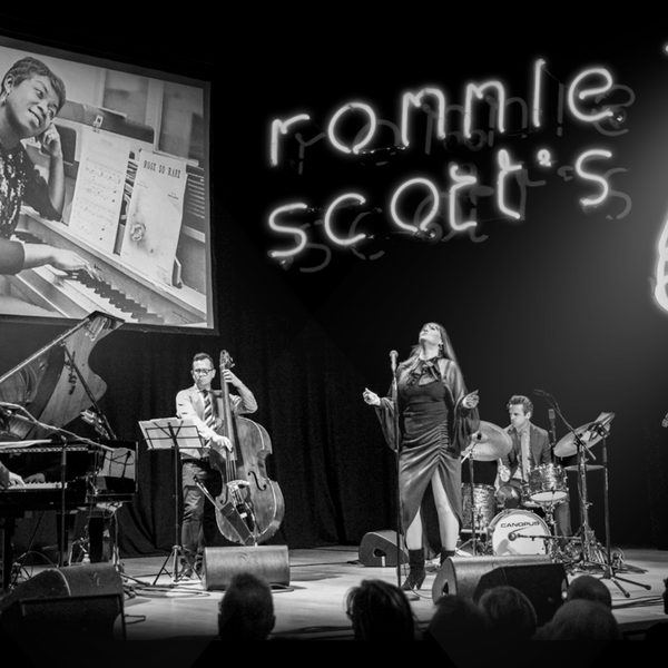 Black and white image of a band performing on stage with Ronnie Scott's logo and a saxophone on the wall