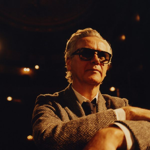 Guy Chambers wearing a grey suit and dark glasses, looking away from the camera with cream hue lighting