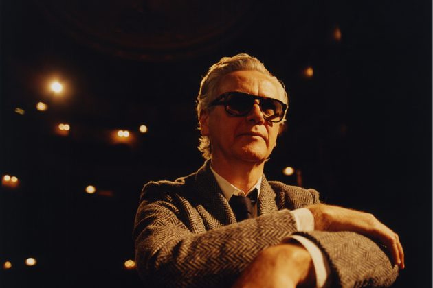 Guy Chambers wearing a grey suit and dark glasses, looking away from the camera with cream hue lighting