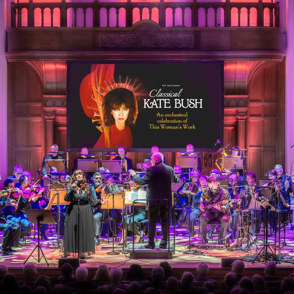 A Kate Bush impersonator wearing a black dress performing on stage with an orchestra and purple lighting