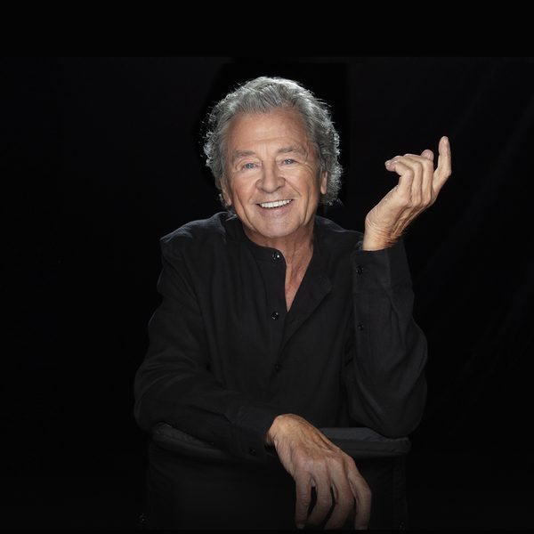 Ian Gillan, a white man with grey hair smiling at the camera with his left hand lifted up, wearing a black shirt in front of a black background
