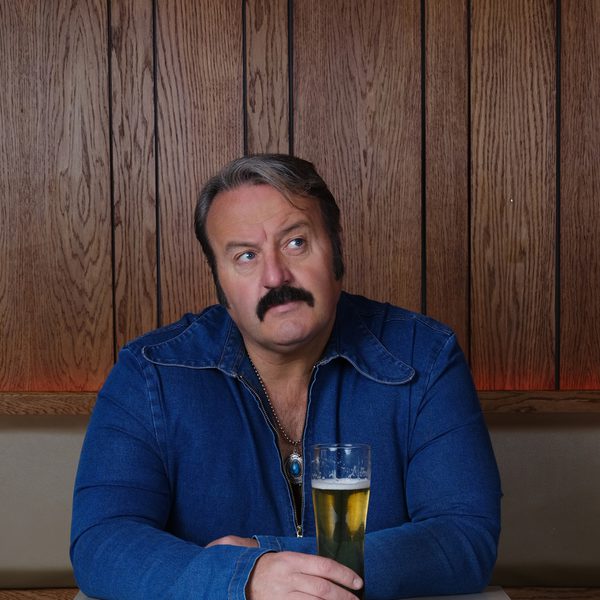 Mike Bubbins, a white man with light hair, a black mustache, wearing a blue denim jacket and holding a drink