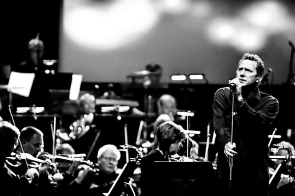 Black and white image of Andy McCluskey, a white man with short hair, singing into a microphone next to orchestra members