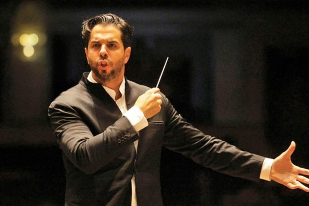 Domingo Hindoyan, a tall Hispanic man with short black hair and beard, wearing a black suit conducting with his arms wide