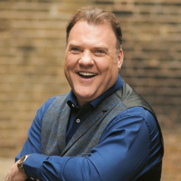 Sir Bryn Terfel, a white man with short brown hair, wearing a blue shirt and waistcoat and leaning over a chair laughing