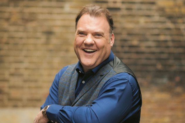 Sir Bryn Terfel, a white man with short brown hair, wearing a blue shirt and waistcoat and leaning over a chair laughing