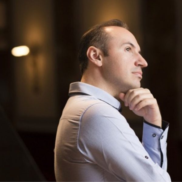 Simon Trpčeski, a white man with short brown hair, wearing a long sleeve white shirt and leaning on his hand looking off to the right
