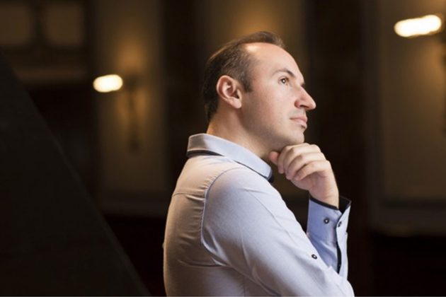 Simon Trpčeski, a white man with short brown hair, wearing a long sleeve white shirt and leaning on his hand looking off to the right