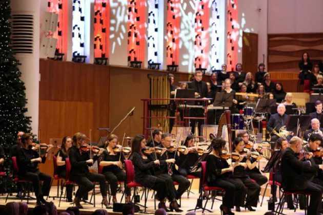 Members of the Orchestra performing in all black on stage, surrounded by Christmas trees and snowflake shaped light projections