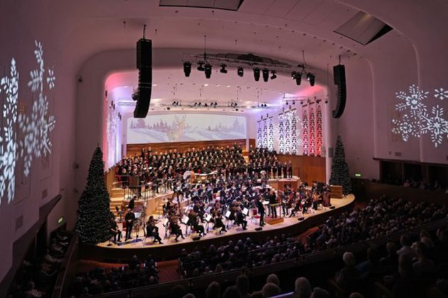 A wide shot of members of the Orchestra performing on stage, surrounded by Christmas trees and snowflake shaped light projections