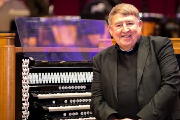 Ian Tracey, a white man with short grey hair, wearing a black suit sat next to an organ