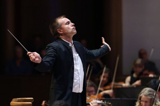 Vasily Petrenko, a tall white man with short brown hair, wearing a black suit conducting the orchestra with his arms outstretched