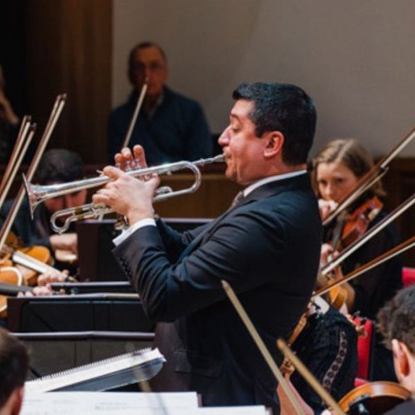 Pacho Flores, a Hispanic man with short black hair, wearing a black suit and playing the trumpet surrounded by violinists