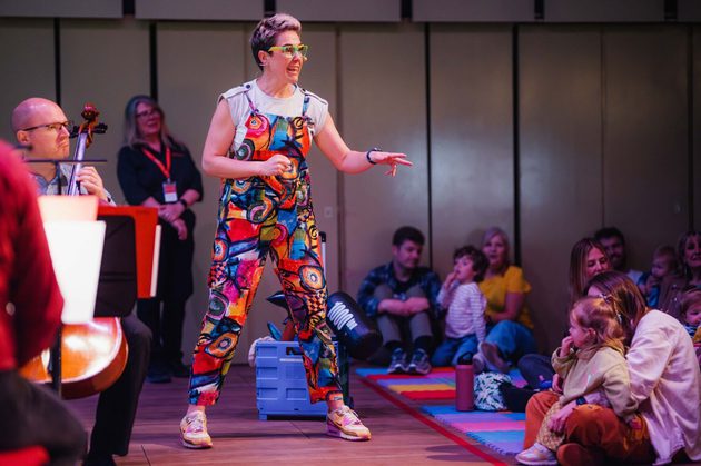 Claire Henry, a white woman with short blonde hair and green glasses, wearing a multicoloured jumpsuit in front of a family audience