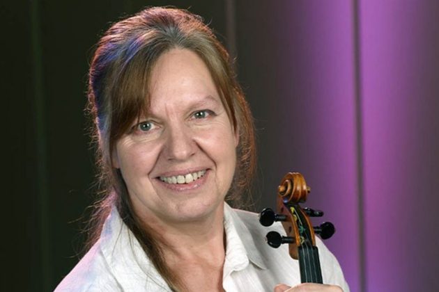 Thelma Handy, a white woman with long brown hair, wearing a white shirt and holding her violin