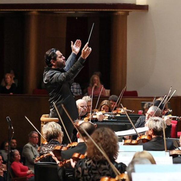 Domingo Hindoyan, a Hispanic man with short black hair, conducting the orchestra from the stand with his arms in the air