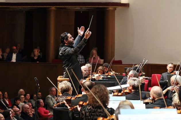 Domingo Hindoyan, a Hispanic man with short black hair, conducting the orchestra from the stand with his arms in the air