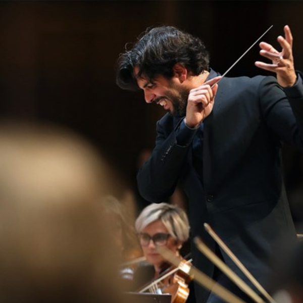 Domingo Hindoyan, a Hispanic man with short black hair, conducting from the stand with his arms in a sweeping motion