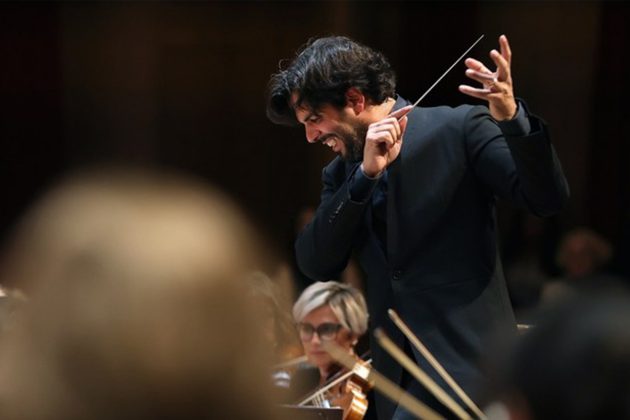 Domingo Hindoyan, a Hispanic man with short black hair, conducting from the stand with his arms in a sweeping motion