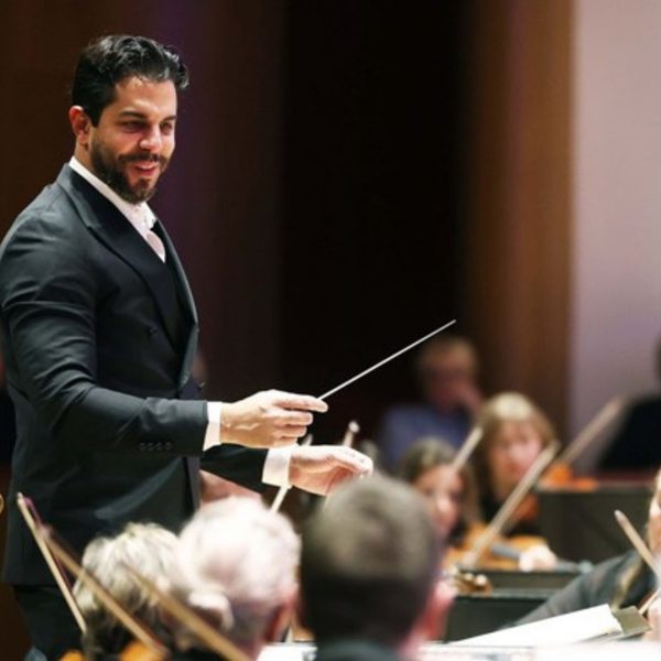 Domingo Hindoyan, a Hispanic man with short black hair, conducting from the stand and smiling at the orchestra