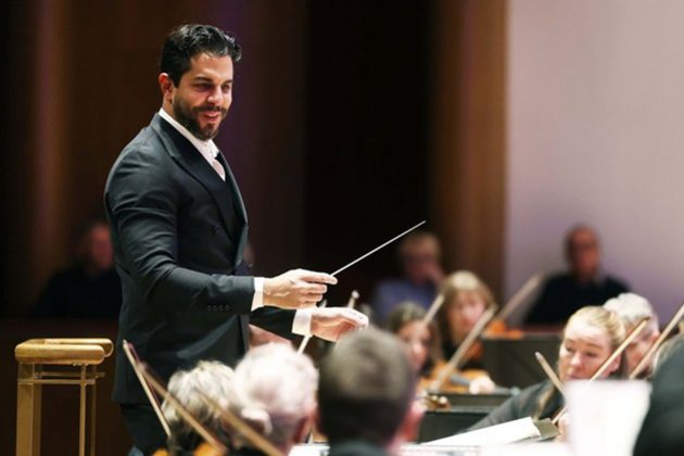 Domingo Hindoyan, a Hispanic man with short black hair, conducting from the stand and smiling at the orchestra