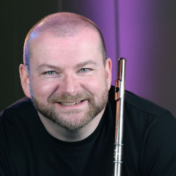 Cormac Henry, a white bald man with a beard, smiling at the camera holding his flute