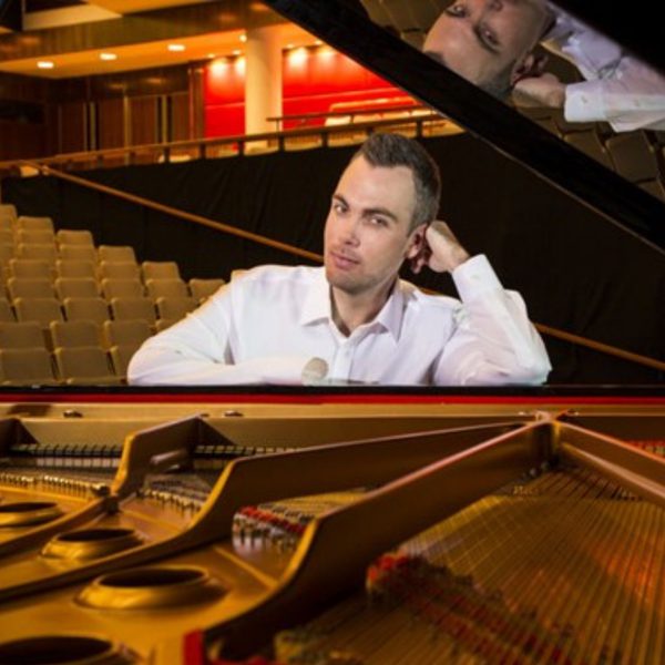 Nicholas McCarthy, a white man with short brown hair, wearing a shirt and leaning on an open piano in an empty auditorium