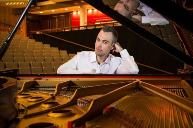 Nicholas McCarthy, a white man with short brown hair, wearing a shirt and leaning on an open piano in an empty auditorium