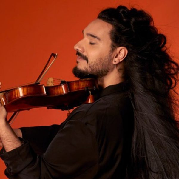 Nemanja Radulović, a Serbian man with long black hair and a beard, playing his violin in front of a bright orange background