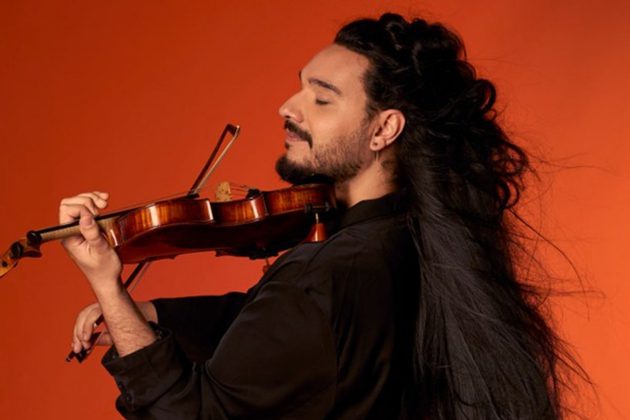 Nemanja Radulović, a Serbian man with long black hair and a beard, playing his violin in front of a bright orange background