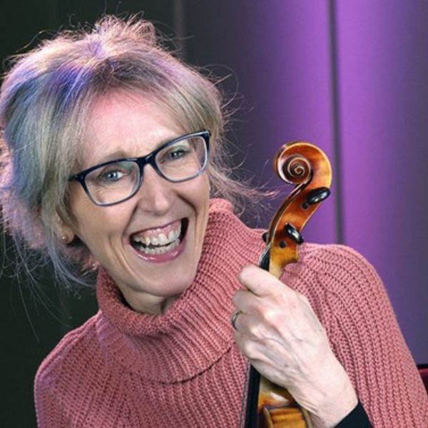 Kate Richardson, a white woman with long grey hair tied up and square glasses, wearing a pink turtleneck and holding her violin