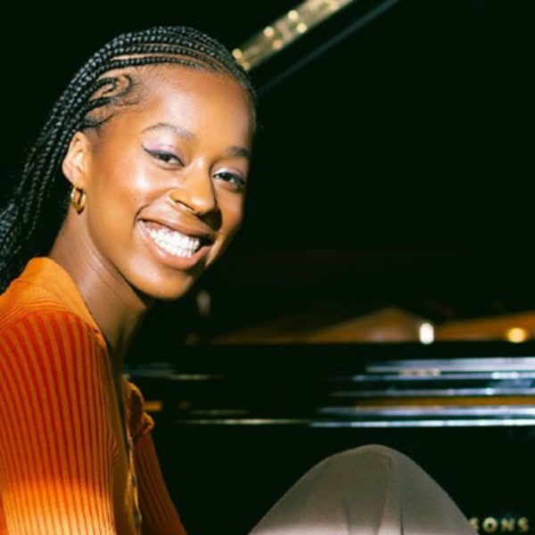 Jeneba Kanneh-Mason, a black woman with long black braids, sat at a piano and wearing an orange top