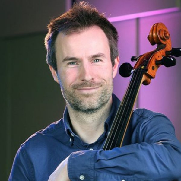 Alexander Holladay, a white man with short grey hair and a beard, wearing a blue shirt and leaning on his cello