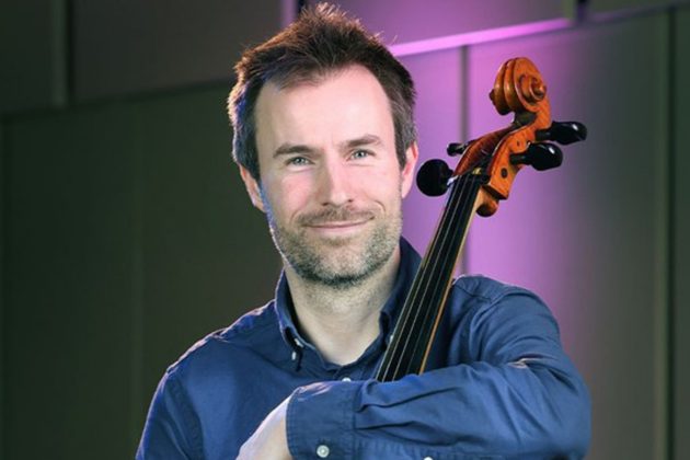 Alexander Holladay, a white man with short grey hair and a beard, wearing a blue shirt and leaning on his cello
