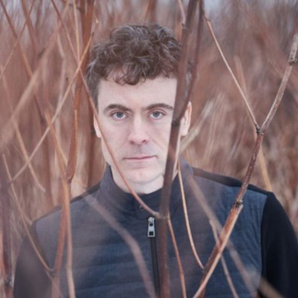 Paul Lewis, a white man with curly grey hair, stood amongst bare trees wearing a black jacket