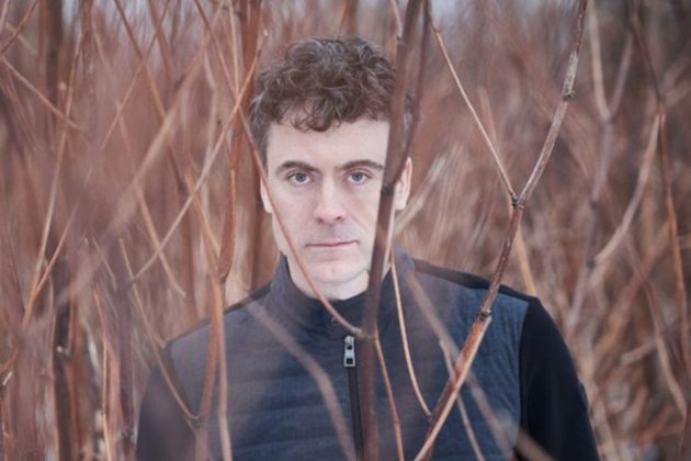 Paul Lewis, a white man with curly grey hair, stood amongst bare trees wearing a black jacket