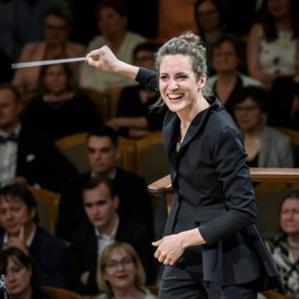 Elena Hron, a white woman with brown hair tied in a bun, wearing all black and conducting from the stand with her arm outstretched