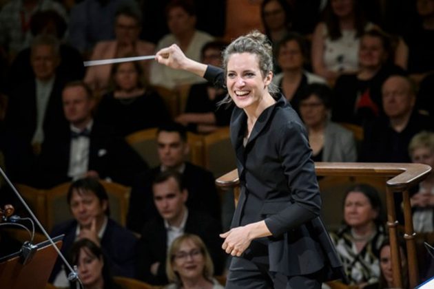 Elena Hron, a white woman with brown hair tied in a bun, wearing all black and conducting from the stand with her arm outstretched