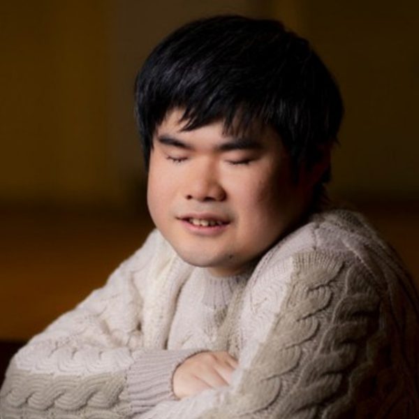 Nobuyuki Tsujii, an Asian man with short black hair, with his arms crossed on a piano wearing a white knitted jumper