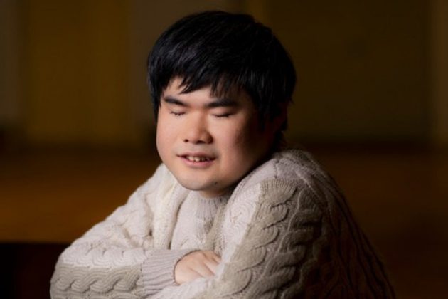Nobuyuki Tsujii, an Asian man with short black hair, with his arms crossed on a piano wearing a white knitted jumper