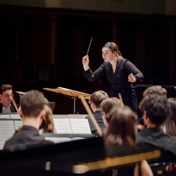 Tess Jackson, a white woman with black hair in a ponytail, wearing all black and conducting an orchestra from the stand