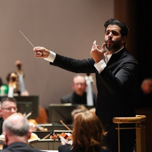 Domingo Hindoyan, a Hispanic man with short black hair and beard, conducting the orchestra from the stand