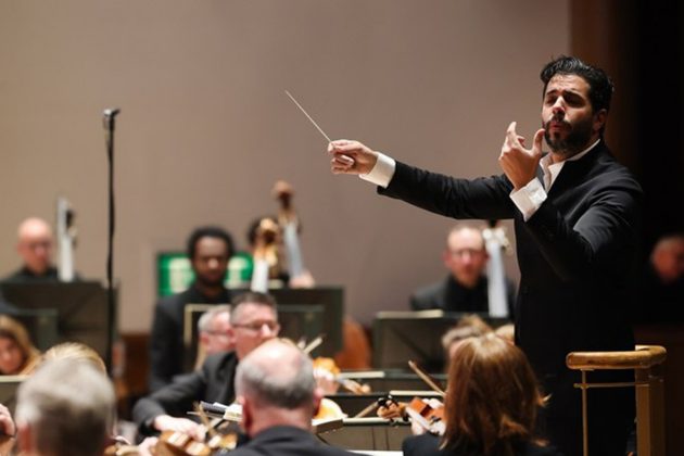 Domingo Hindoyan, a Hispanic man with short black hair and beard, conducting the orchestra from the stand