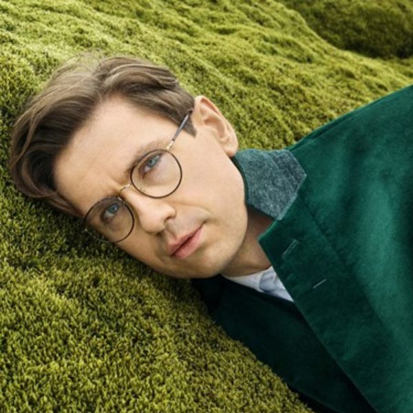 Víkingur Ólafsson, a white man with short brown hair and round glasses, wearing a green velvet jacket and lying on green grass