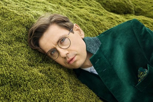 Víkingur Ólafsson, a white man with short brown hair and round glasses, wearing a green velvet jacket and lying on green grass