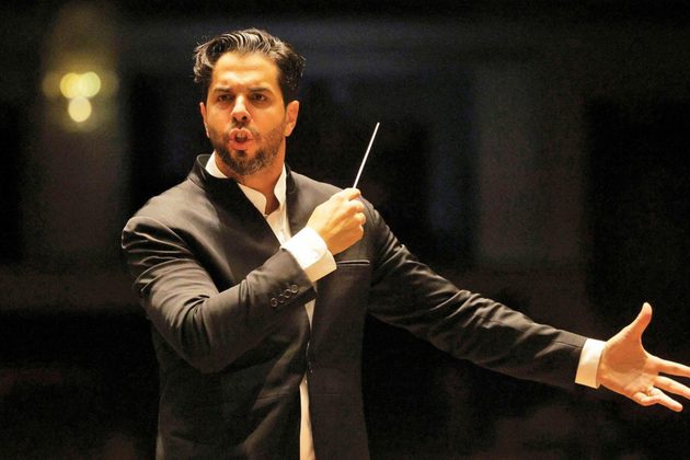 Domingo Hindoyan, a tall Hispanic man with short black hair and beard, wearing a black suit conducting with his arms wide